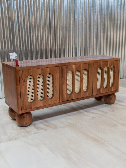 A mid-century modern wooden sideboard cabinet with three doors featuring woven cane panels, round wooden legs, and a warm medium-brown finish, set against a corrugated metal wall.