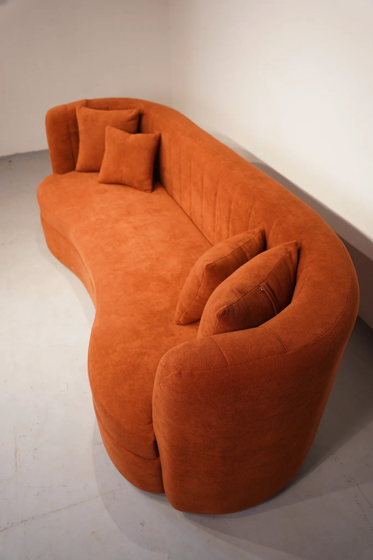 A modern, curved orange velvet sofa with matching throw pillows, photographed in a minimalist studio setting with a white wall and light gray floor.