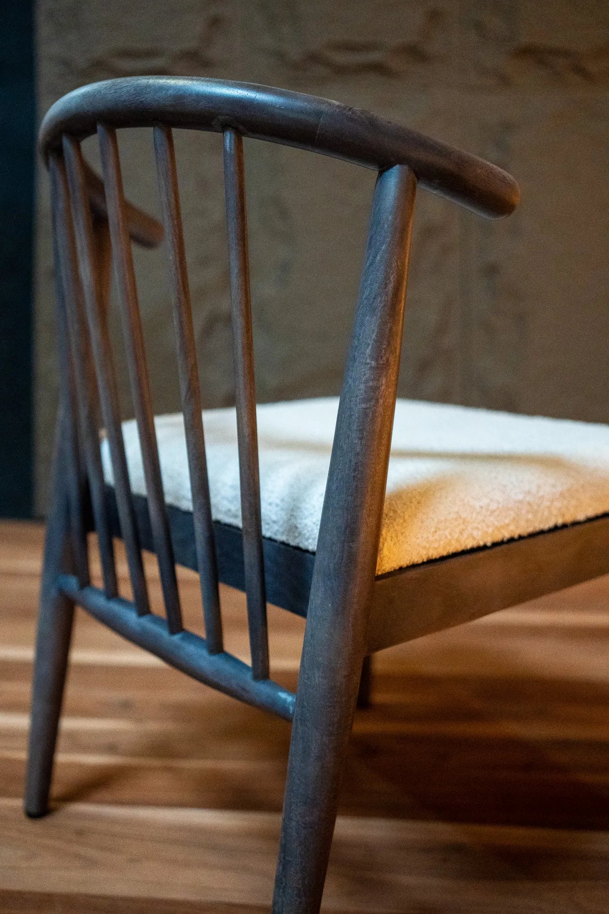 A close-up of a mid-century modern wooden chair with a curved backrest, dark weathered gray-brown wood frame, and a cream-colored textured seat cushion, set on a wooden floor against a stone wall.