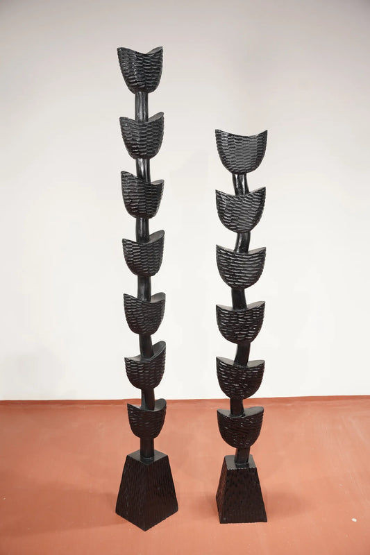 Two tall black sculptural wall or floor pieces with stacked curved leaf-like forms on tapered rectangular bases, photographed indoors against a plain light wall and terracotta floor.
