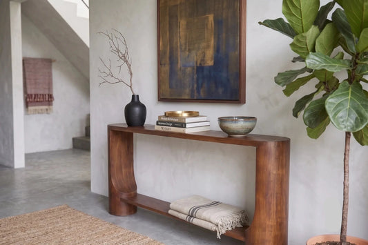 A modern wooden console table in a styled entryway, styled with books, a black vase, a bowl, and a folded throw blanket. A large potted fiddle-leaf fig plant stands to the right, and a textured abstract painting hangs on the wall behind the table.
