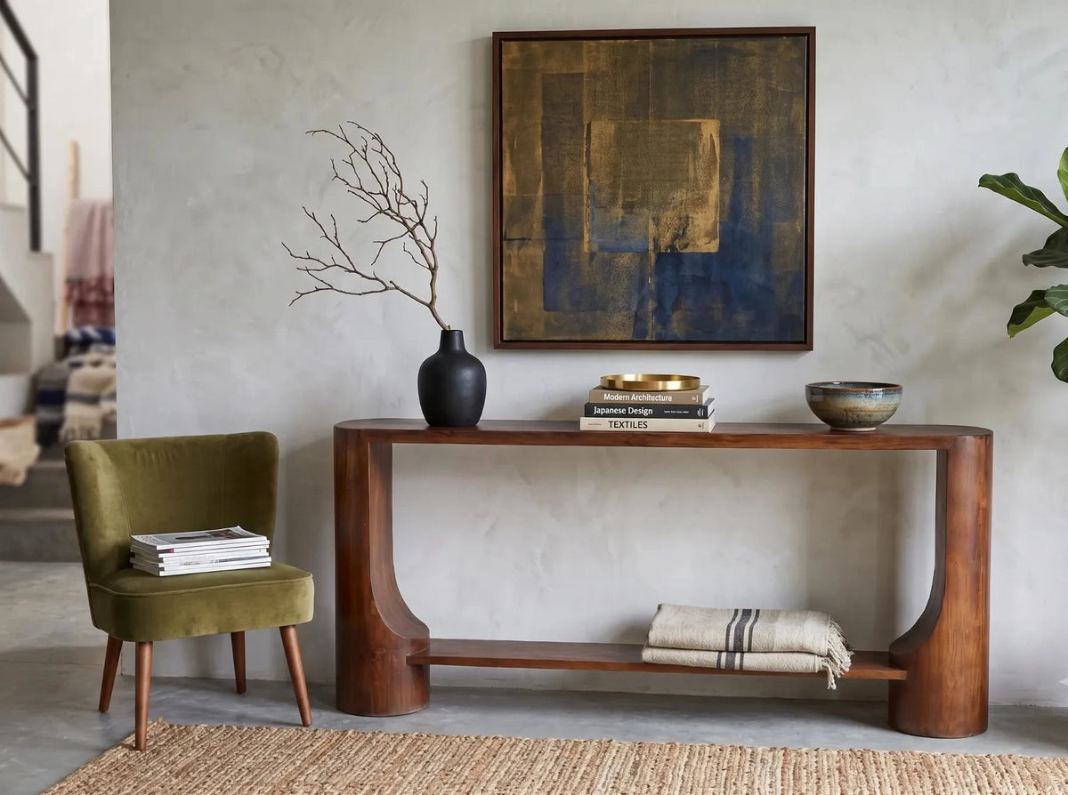 A mid-century modern wooden console table styled in a living room setting with a green armchair, a potted fiddle-leaf fig plant, a textured abstract painting, and decorative accessories.