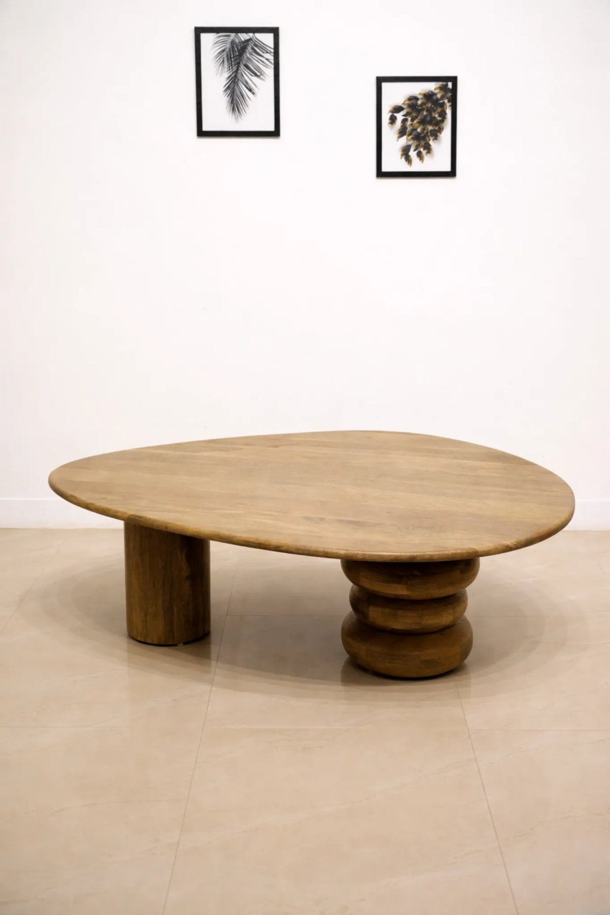 An organic-shaped wooden coffee table with a light natural wood top and a thick, stacked cylindrical base, set against a minimalist white wall and beige tiled floor.