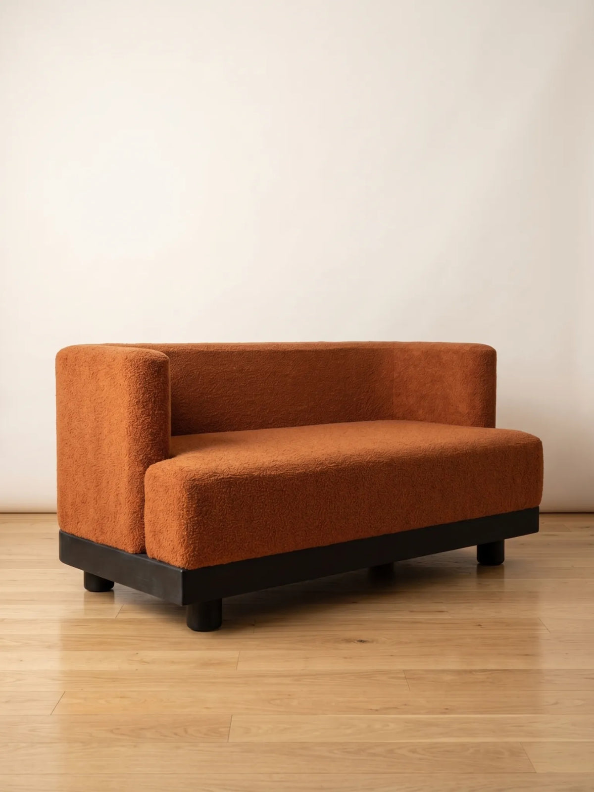 A modern low-profile loveseat with a textured rust-orange bouclé fabric and a dark wood base, set against a plain off-white wall and light wood floor.