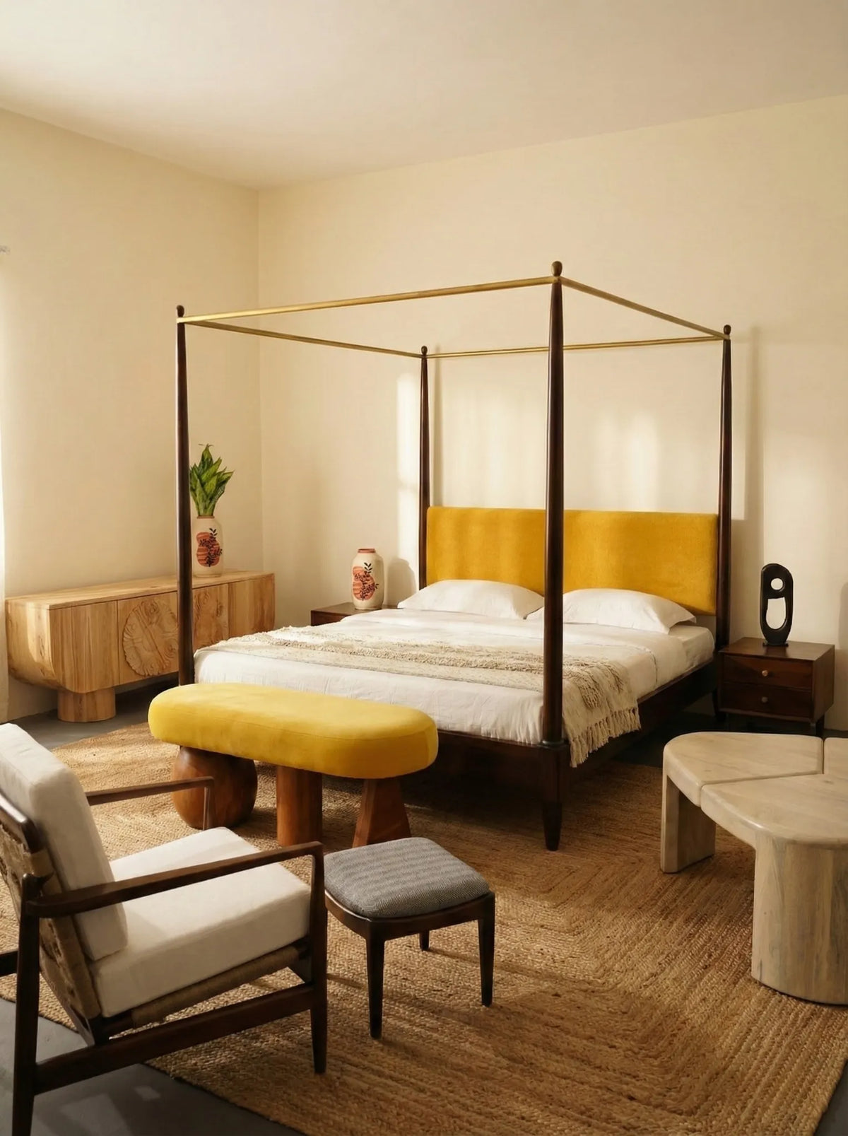 A modern bedroom featuring a four-poster canopy bed with a mustard yellow upholstered headboard and matching yellow bench, with a teak wood accent armchair with rope weave back, along with wooden upholstered stool, a modern classic wooden coffee table, a sideboard/credenza with sustainable papermache vase decor and wooden bedside with wooden decor items all styled by Meraki Arts set in a warm neutral-toned room with a wooden floor and beige walls.