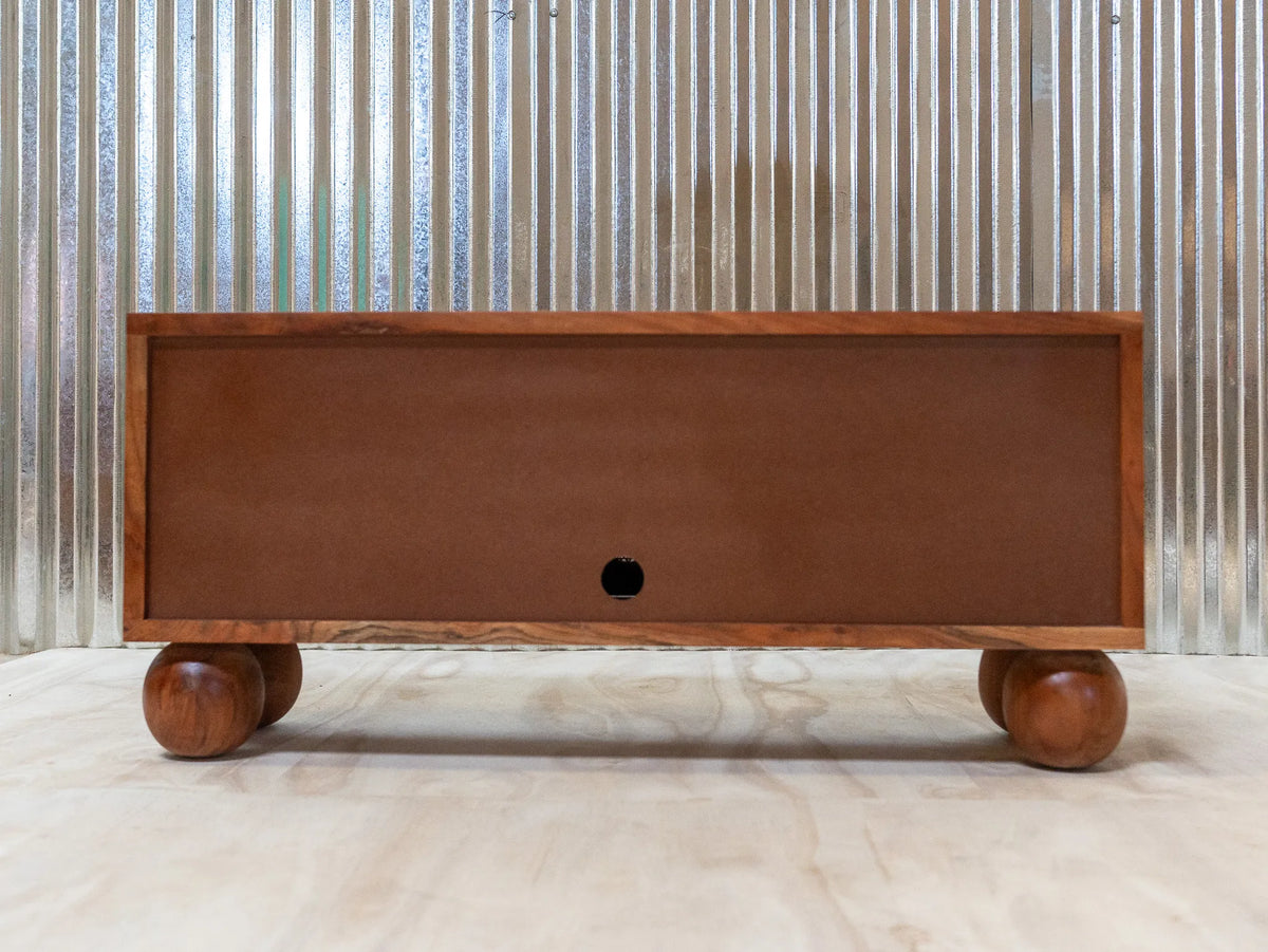 A mid-century modern wooden storage chest with a single drawer, featuring a warm brown wood-grain finish and round wooden feet, set against a corrugated metal wall.