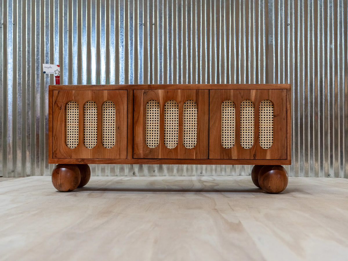 A mid-century modern wooden sideboard cabinet with woven cane panels on the front, resting on round wooden feet against a corrugated metal wall.