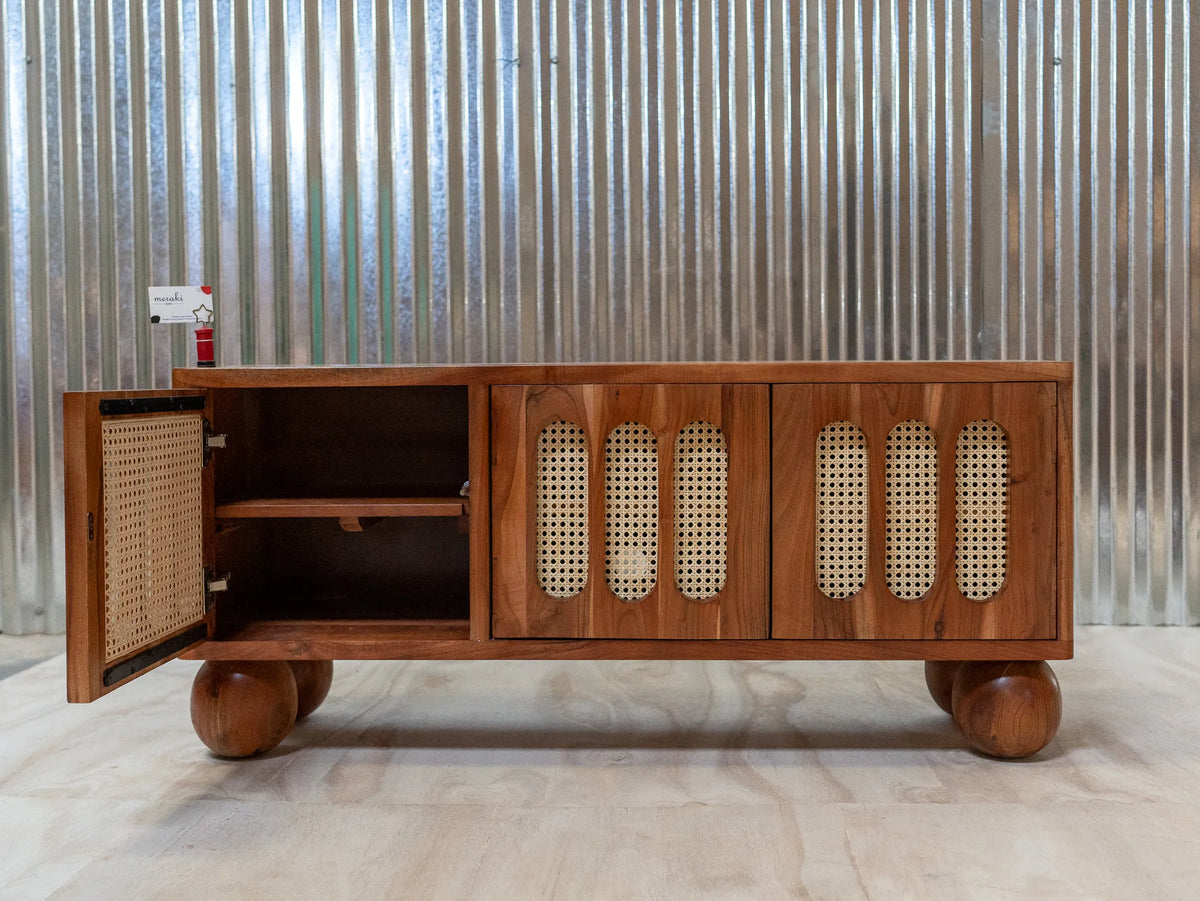 A mid-century modern wooden sideboard cabinet with a warm medium-brown finish, featuring three doors with oval cane inserts and an open side cabinet showing a shelf. The cabinet stands on round wooden feet against a corrugated metal wall.