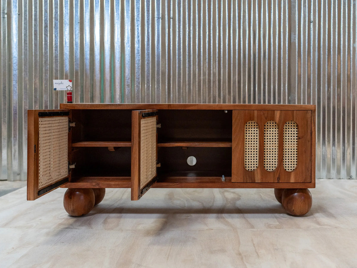 A mid-century modern wooden media console with open doors, woven cane panel doors, and round wooden feet, set against a corrugated metal wall.