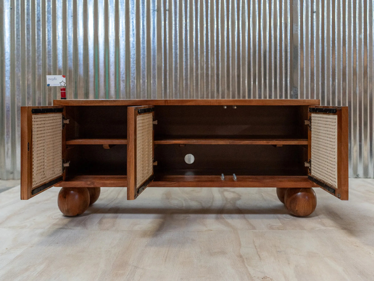 A mid-century modern wooden media console with open cane-paneled doors, shown on a light wood floor against a corrugated metal wall.