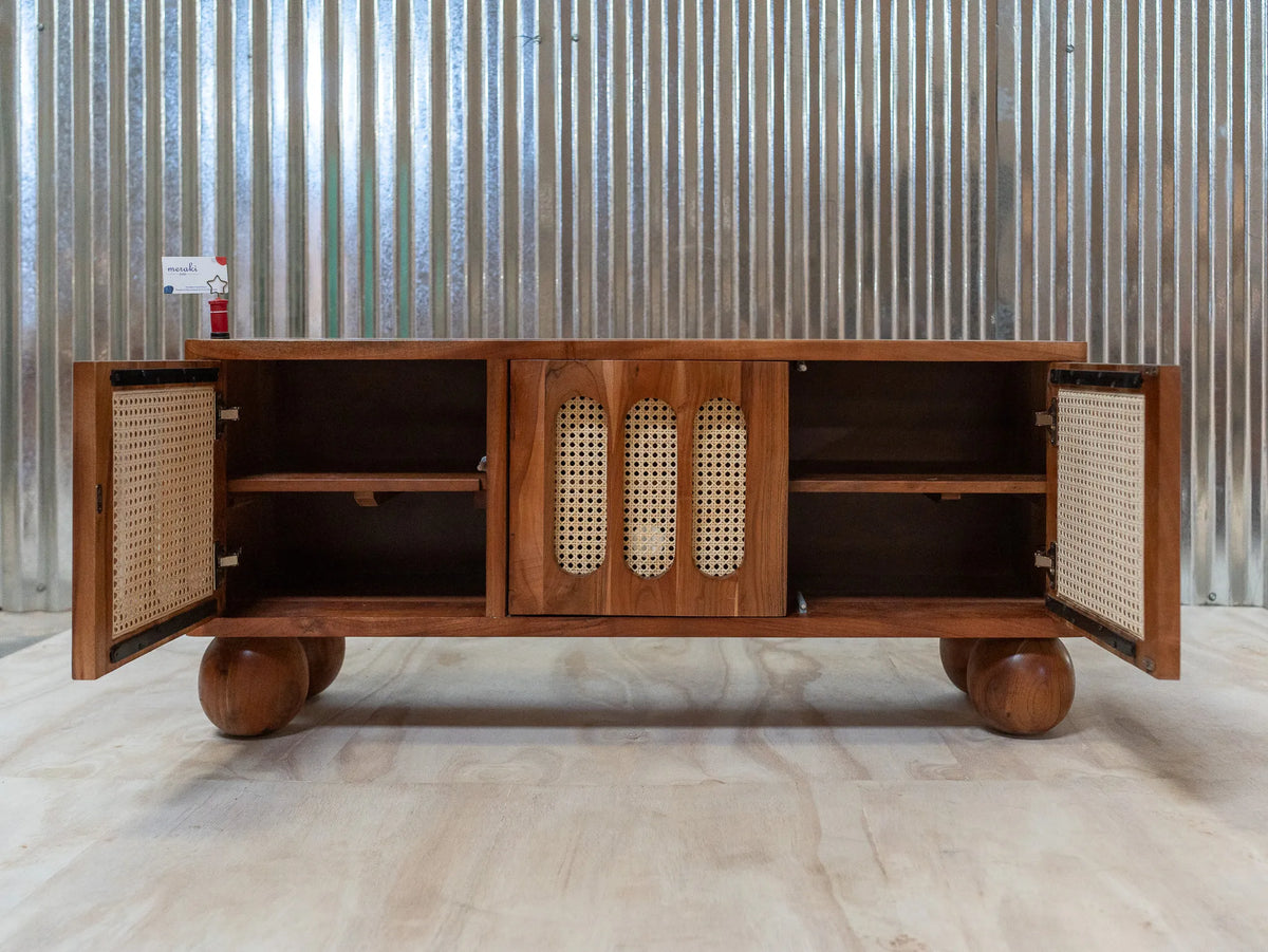 A mid-century modern wooden media console with open cane-paneled doors and round wooden feet, set against a corrugated metal wall.