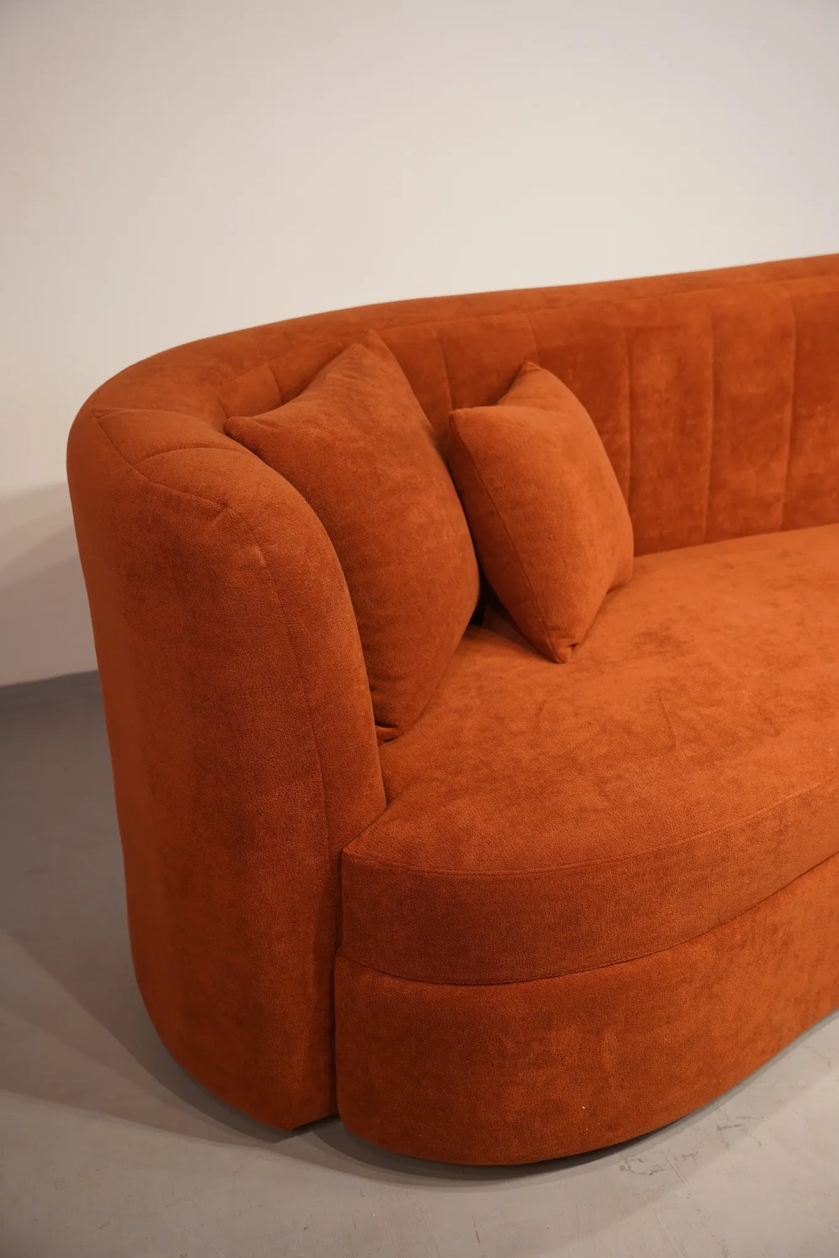 A curved orange velvet sofa with two matching throw pillows, photographed in a minimalist studio setting with a white wall and gray floor.