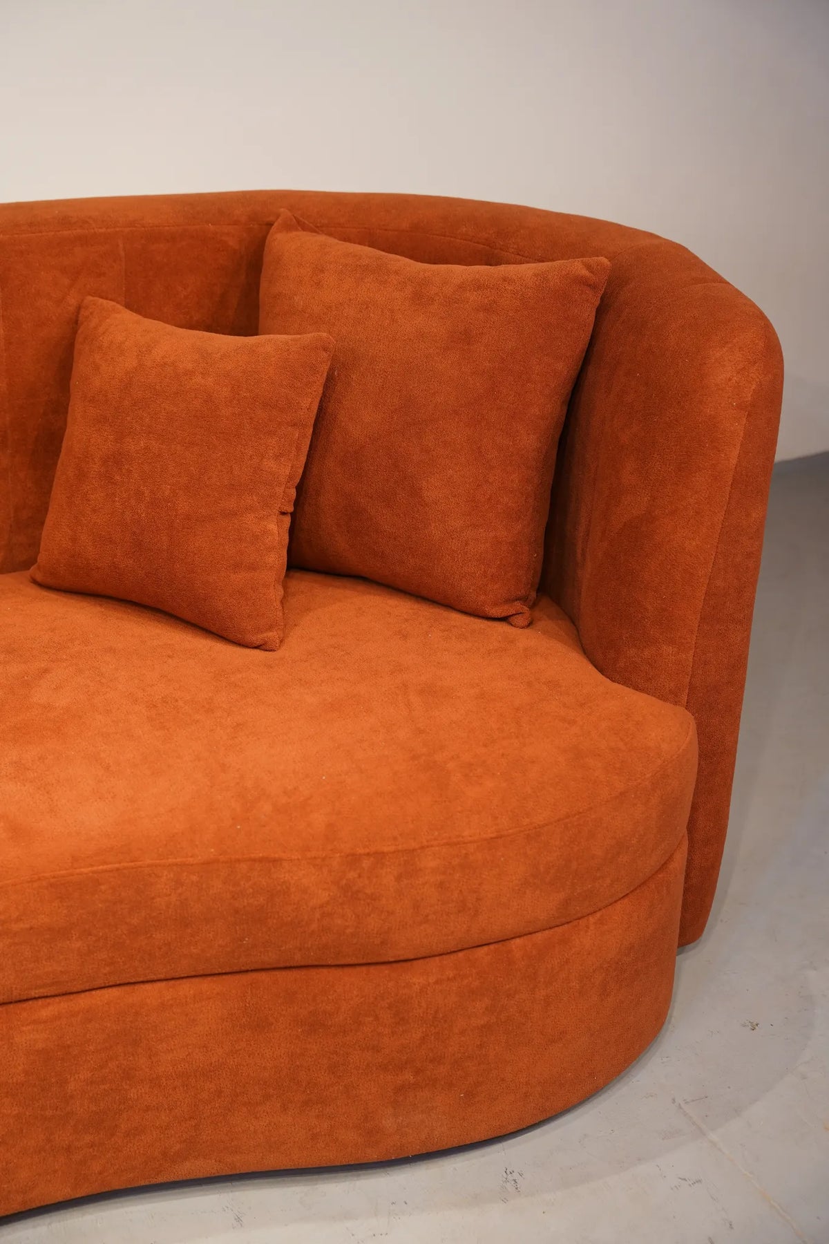 A curved orange upholstered sofa with two matching square throw pillows, shown in a minimalist studio setting with a light gray floor and wall.