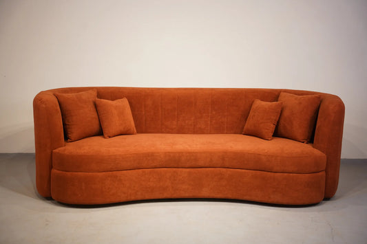 A curved orange-brown velvet sofa with four matching throw pillows, photographed against a plain off-white studio background.
