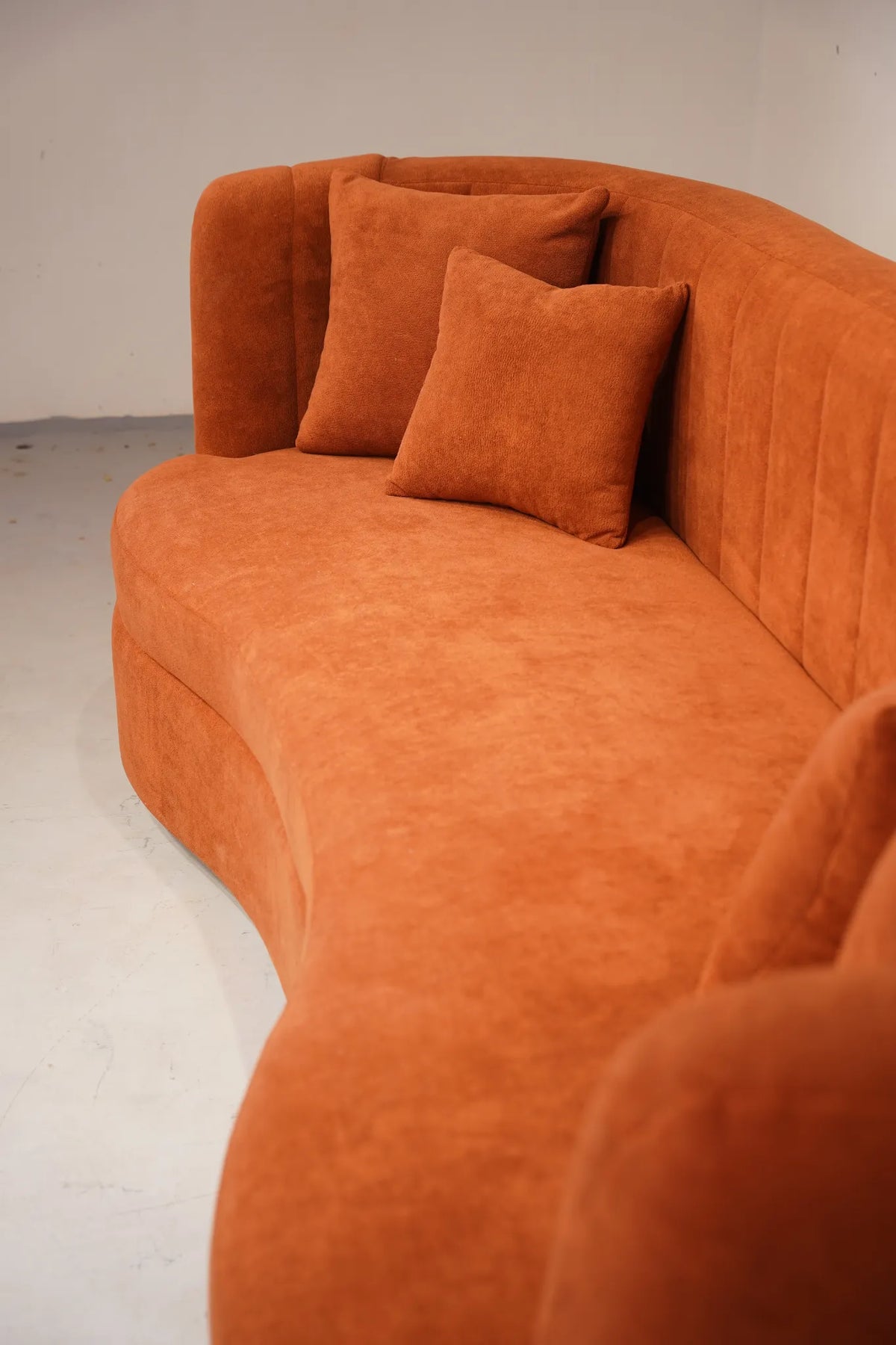 A curved orange velvet sofa with matching throw pillows, photographed in a minimalist studio setting with a light gray floor and wall.