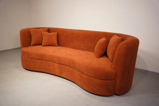 A curved orange velvet sofa with matching throw pillows, photographed in a minimalist studio setting with a white wall and gray floor.
