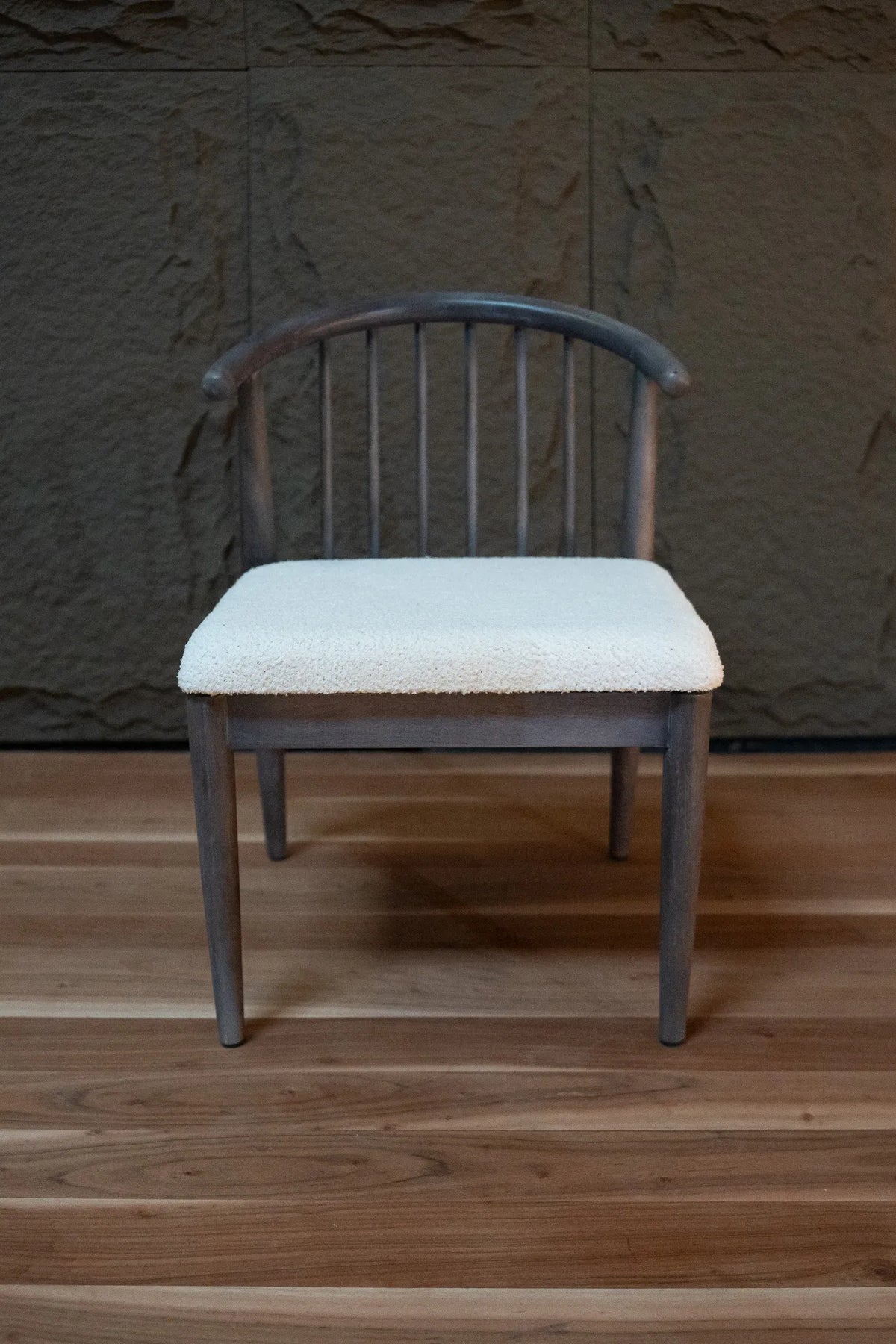 A wooden dining chair with a curved backrest and a cream-colored upholstered seat cushion, set against a dark textured wall and a wooden floor.