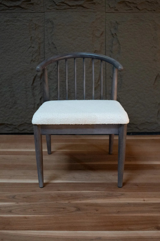 A wooden dining chair with a curved backrest and a cream-colored upholstered seat cushion, set against a dark textured wall and a wooden floor.