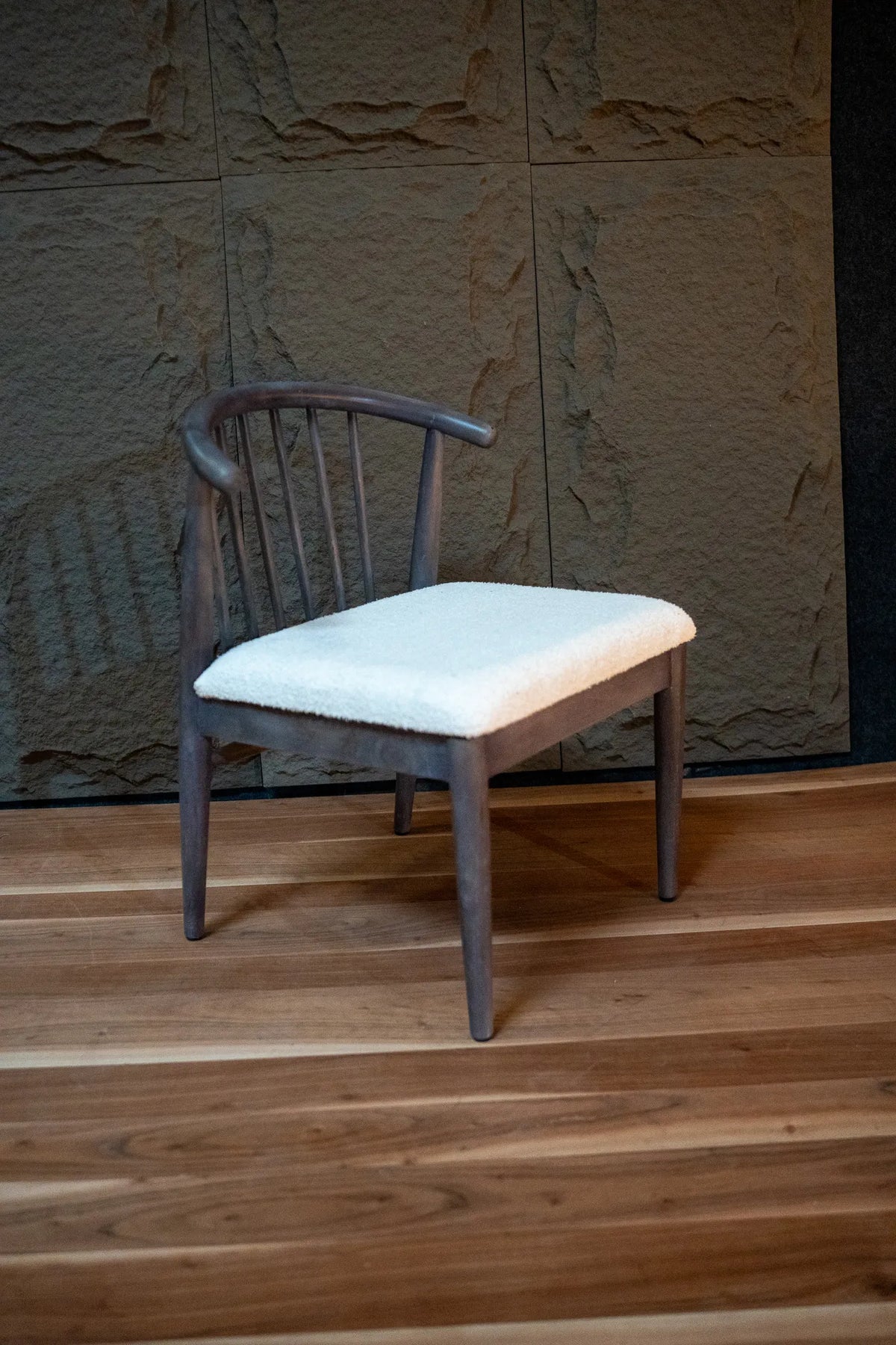 A wooden dining chair with a curved backrest and a white upholstered seat cushion, placed on a wooden floor against a textured stone wall.