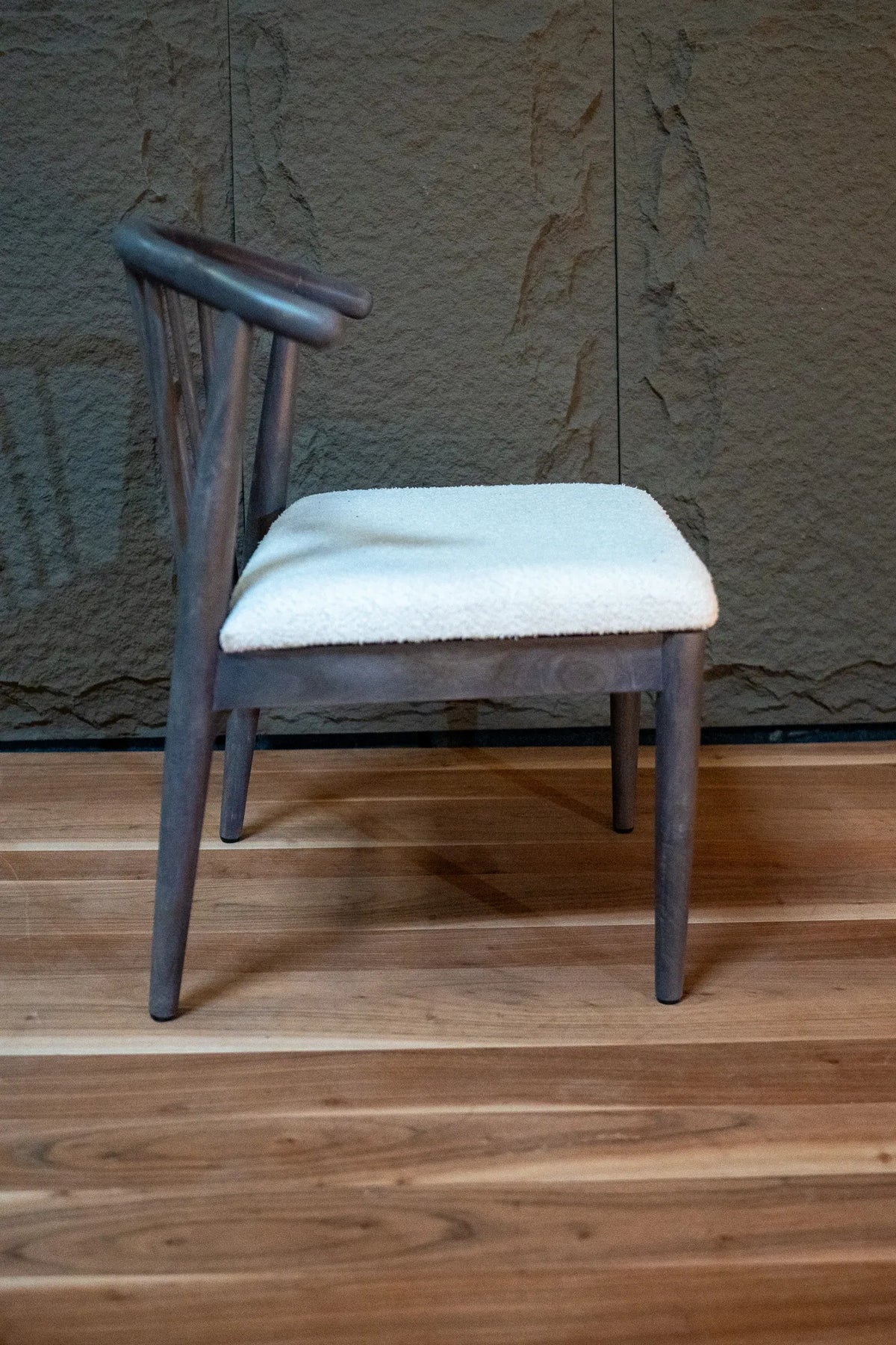 A mid-century modern wooden dining chair with a dark brown frame and a light beige textured seat cushion, set against a rustic stone wall and wooden floor.