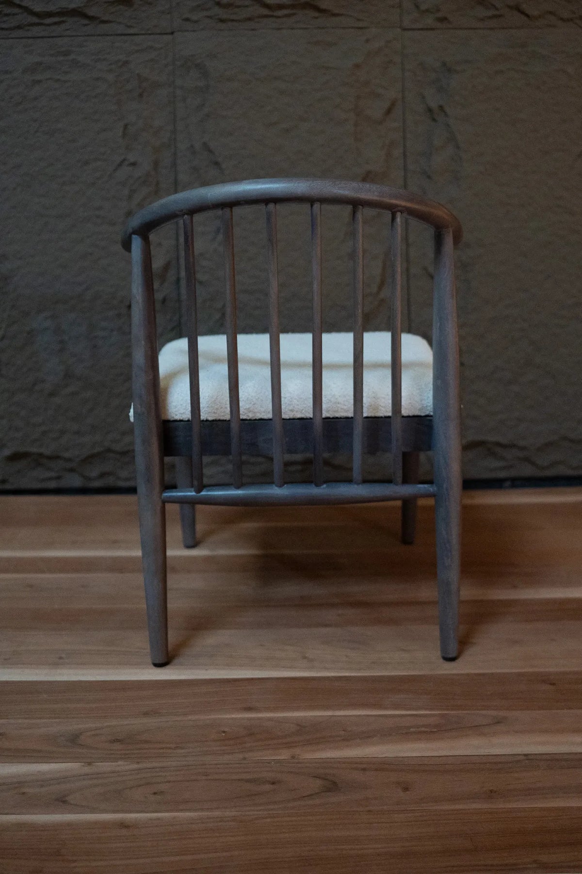 A wooden spindle-back chair with a gray-brown finish and a cream-colored seat cushion, set against a textured stone wall and wooden floor.