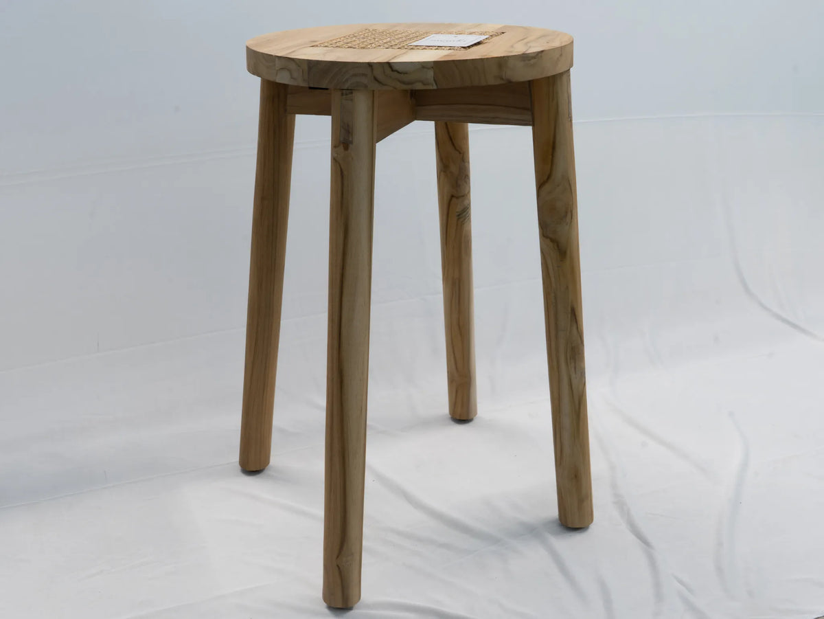 A round wooden stool with four tapered legs and a woven cane seat, shown against a white fabric backdrop.