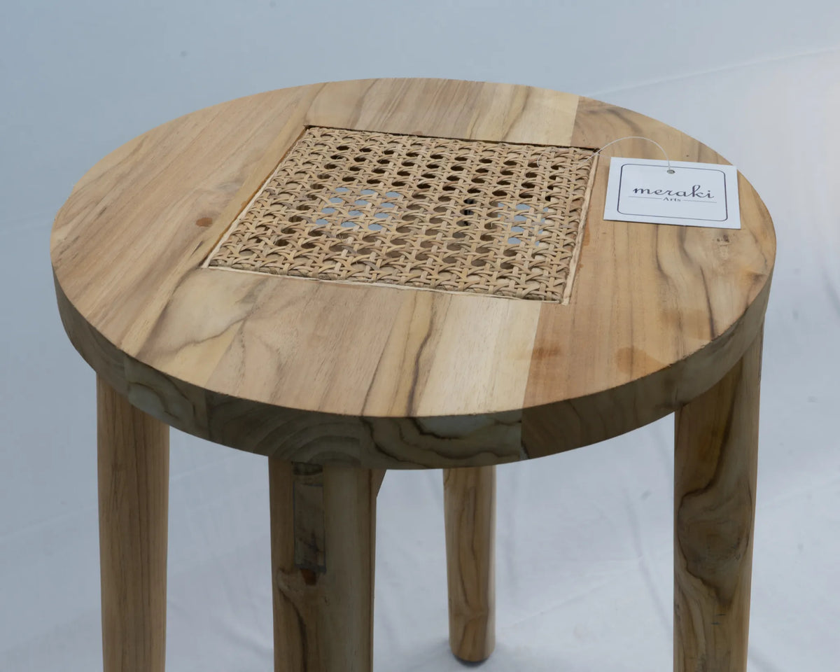 A round teak wooden table with a natural light wood finish, featuring a woven cane or rattan panel in the centre and four sturdy legs. A small white tag with the brand name 'Meraki Arts' is attached to the tabletop.