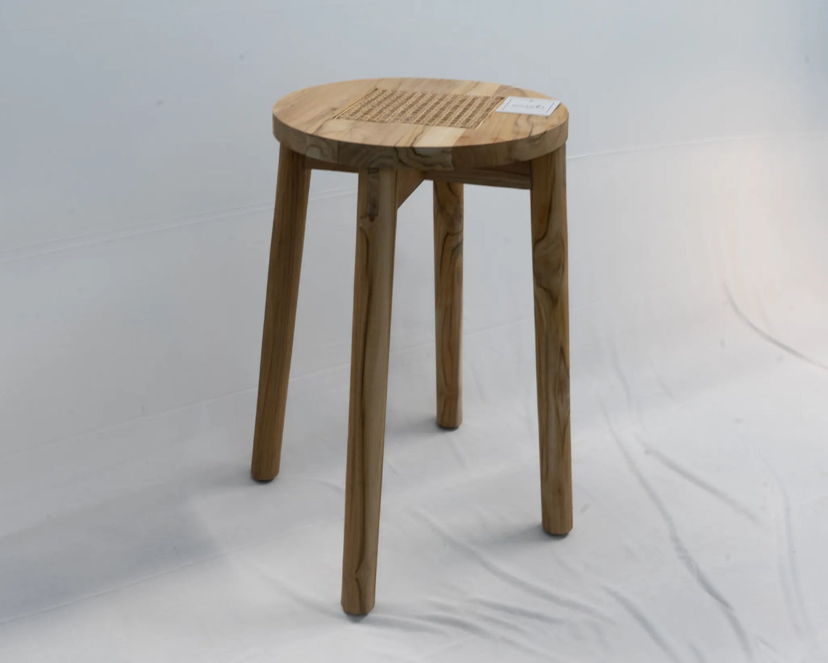 A small round teak wooden stool with four tapered legs and a slatted topwith rattan cane detail, photographed against a plain light gray studio backdrop.