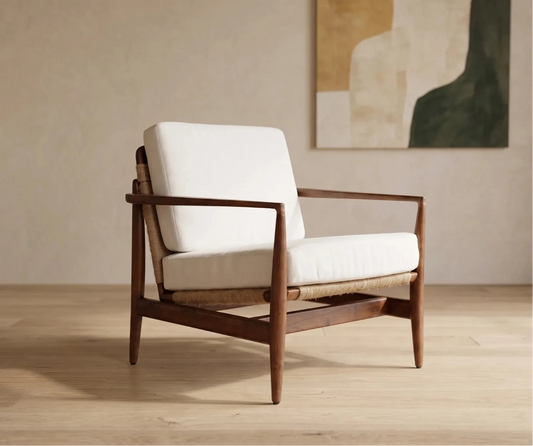 A mid-century modern armchair with a white cushioned seat and backrest, supported by a dark brown wooden frame and a woven cane base, set in a minimalist room with a beige wall and abstract painting.