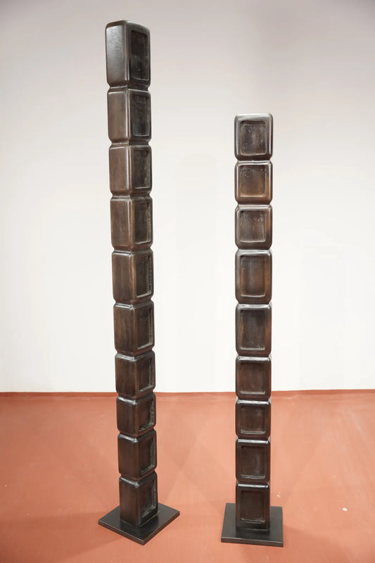 Two dark brown wooden block towers with square bases, standing upright against a plain off-white wall and terracotta-colored floor.