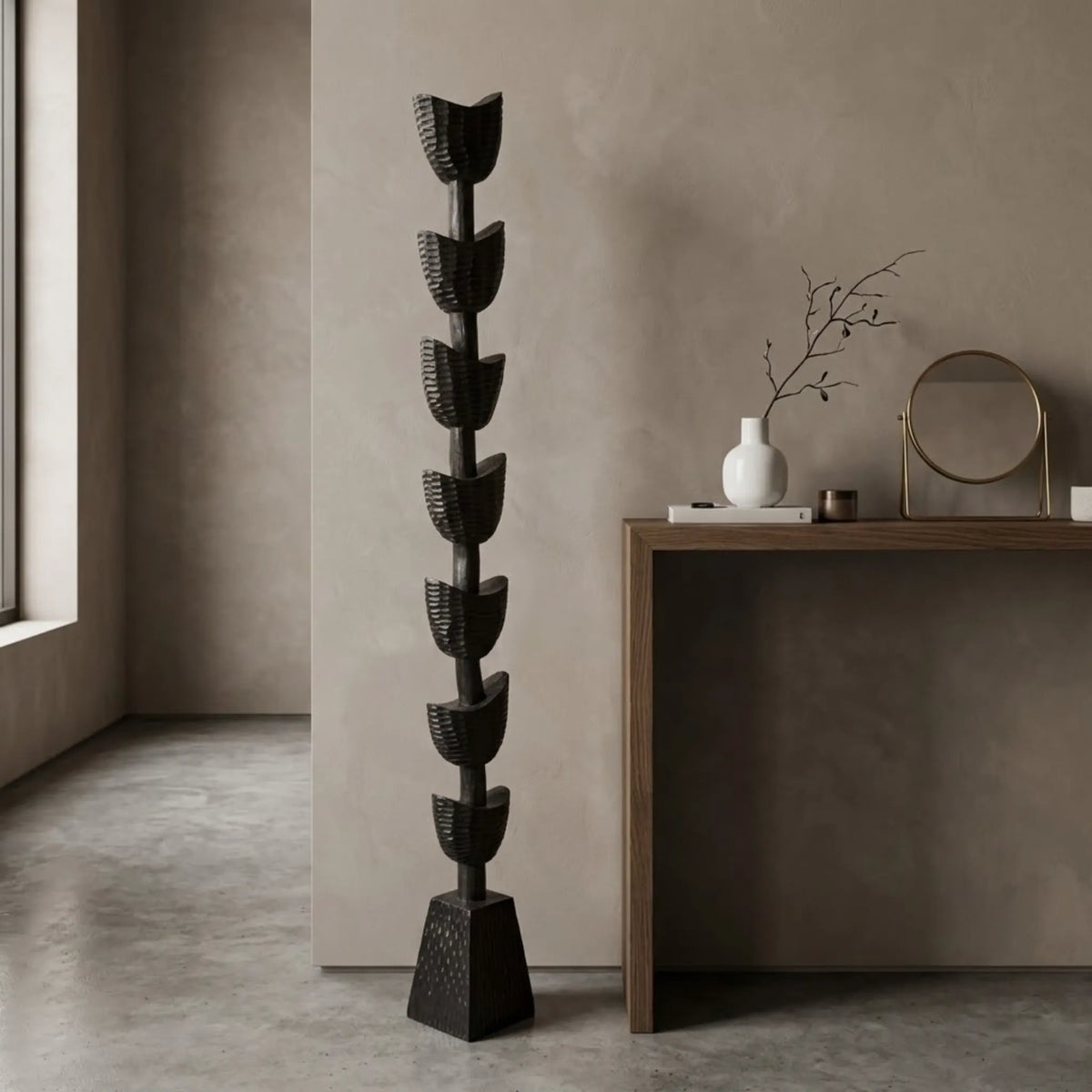 A tall, sculptural black floor sculpture cum totem with a tiered, leaf-like design stands in a minimalist room next to a wooden console table with neutral decor.