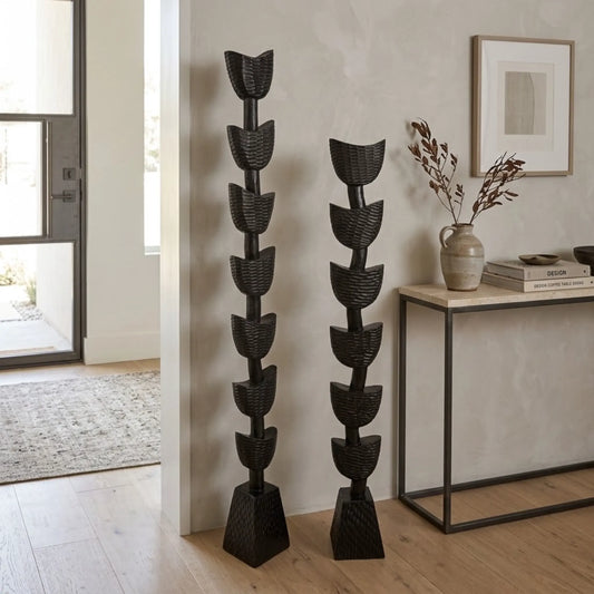 Two tall black woven decorative floor sculptures or totems with stacked leaf-like shapes, displayed in a modern interior next to a console table.