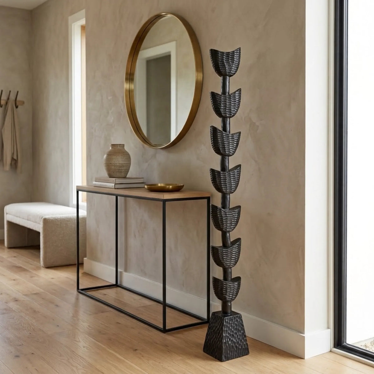 A tall, sculptural black hand carved totem stands in a modern entryway next to a wood console table, round gold-framed mirror, and a light upholstered bench.