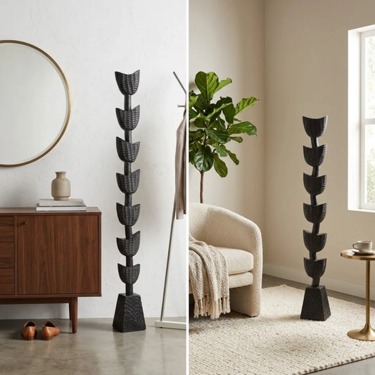 A tall black decorative floor sculpture or totem in 2 sizes with a stacked, leaf-like tiered design stands in a split interior scene: on the left beside a wooden cabinet and mirror, and on the right beside a cream chair, plant, rug, and side table.