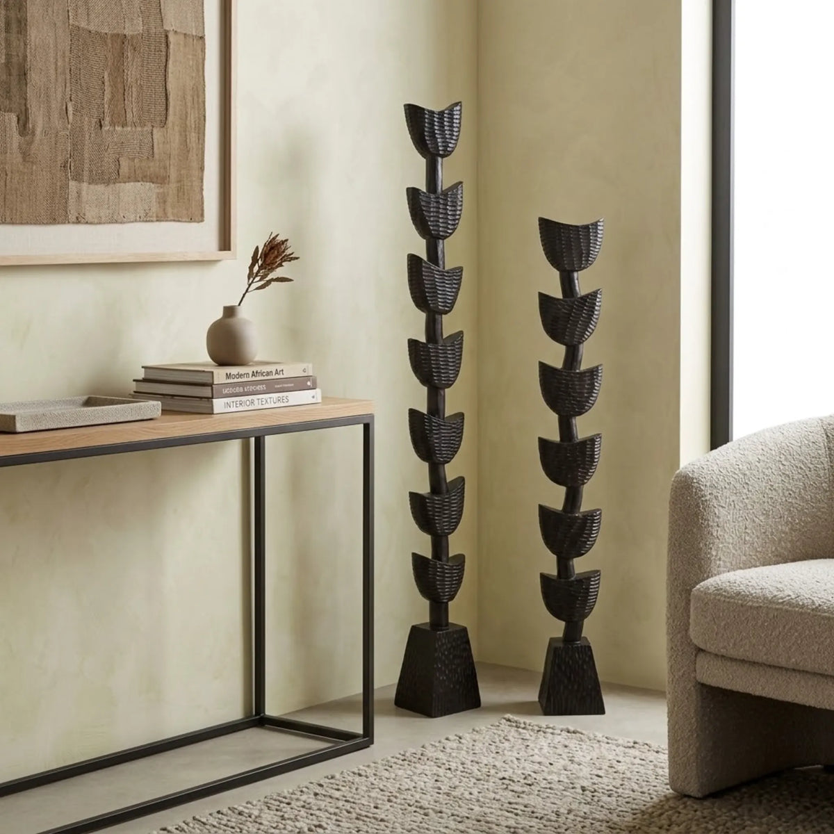 Two tall black woven decorative floor sculptures with stacked leaf-like shapes, placed in a styled interior beside a console table and textured chair.