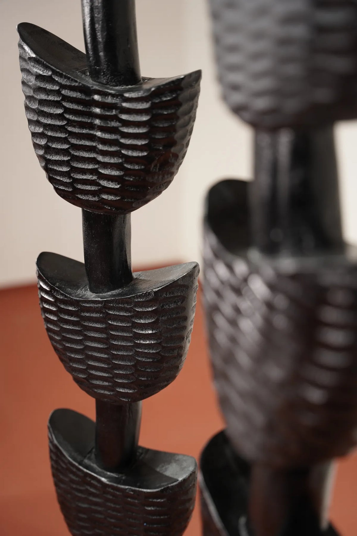 Close-up of a dark carved wooden sculpture with textured, leaf-like or shell-like segments on vertical posts, set against a softly blurred indoor background.