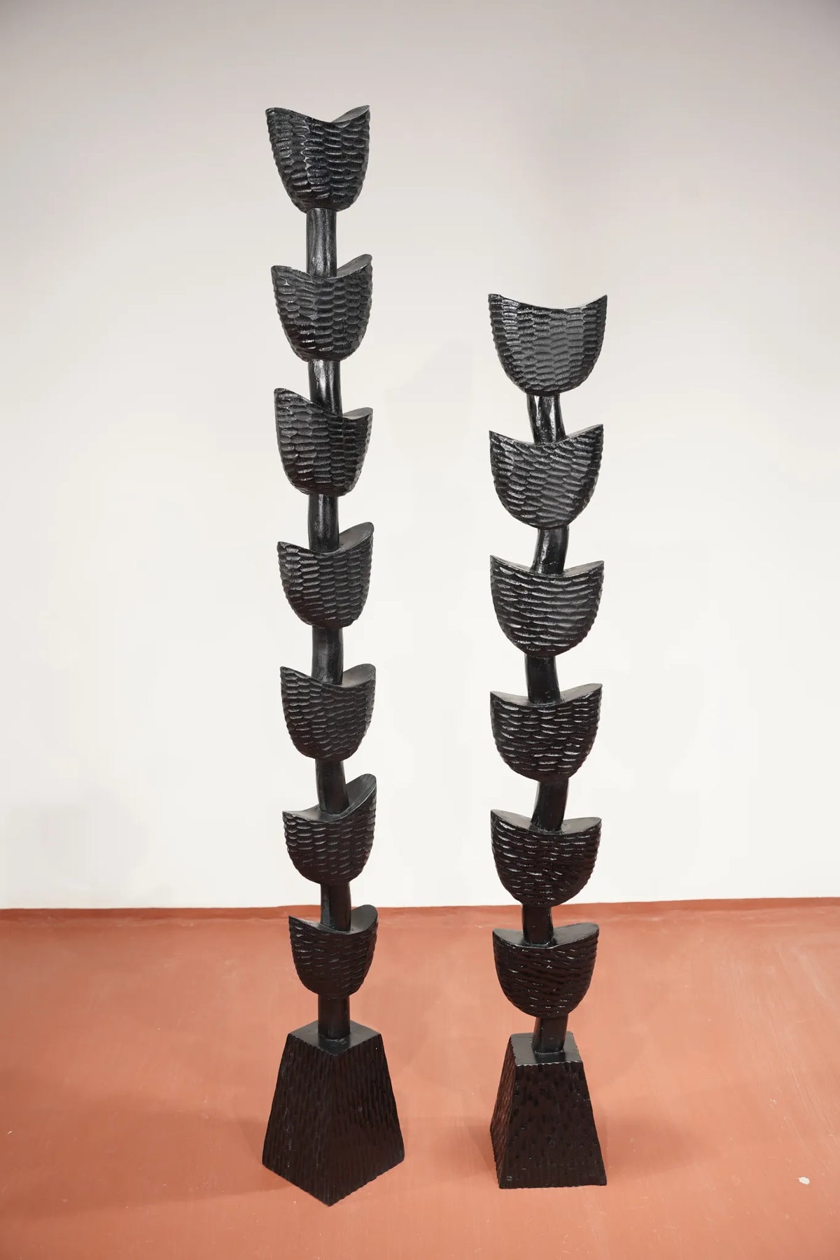 Two tall black sculptural wall or floor pieces with stacked curved leaf-like forms on tapered rectangular bases, photographed indoors against a plain light wall and terracotta floor.