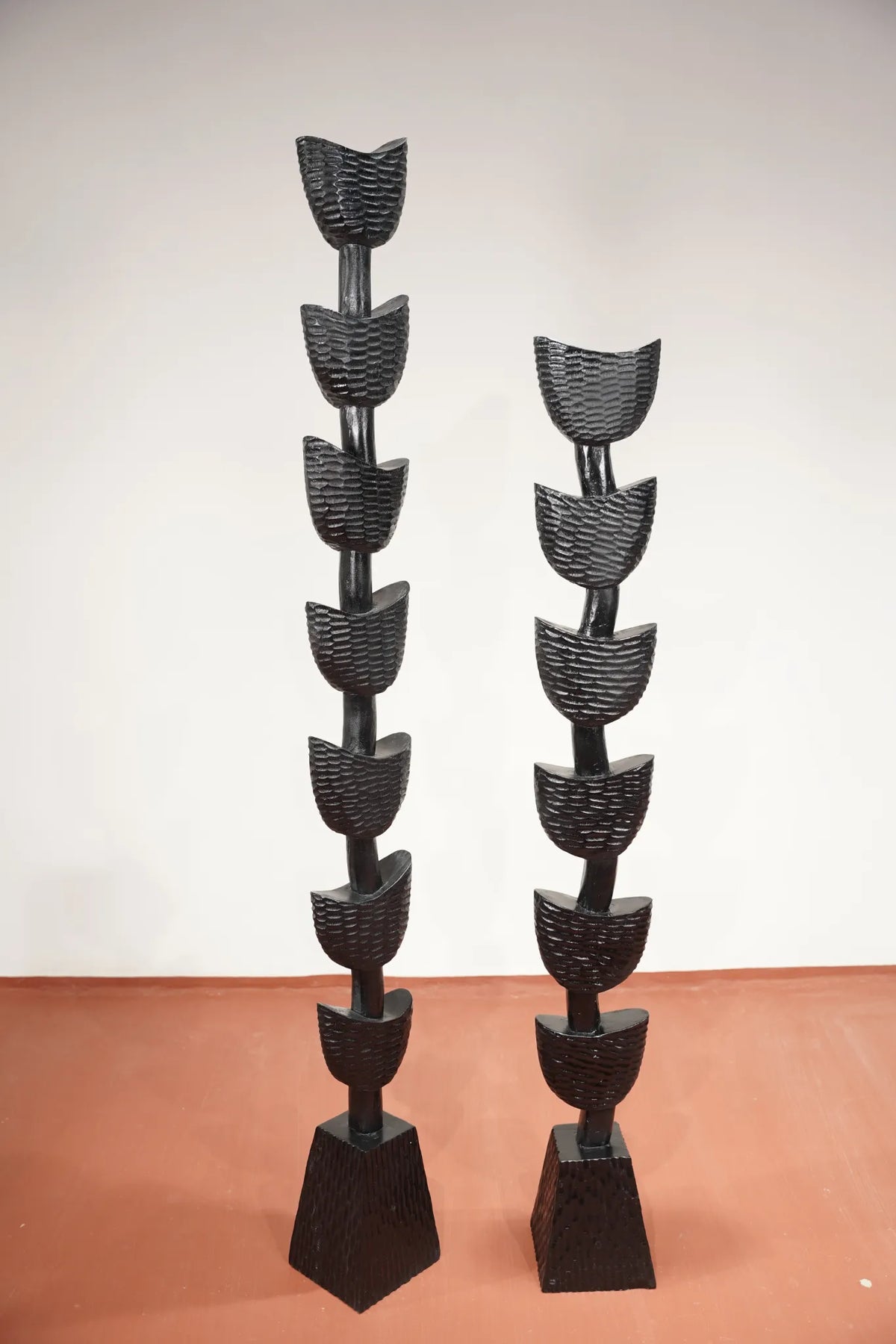 Two tall black sculptural floor pieces with stacked curved leaf-like forms on a terracotta-colored floor against a plain off-white wall.