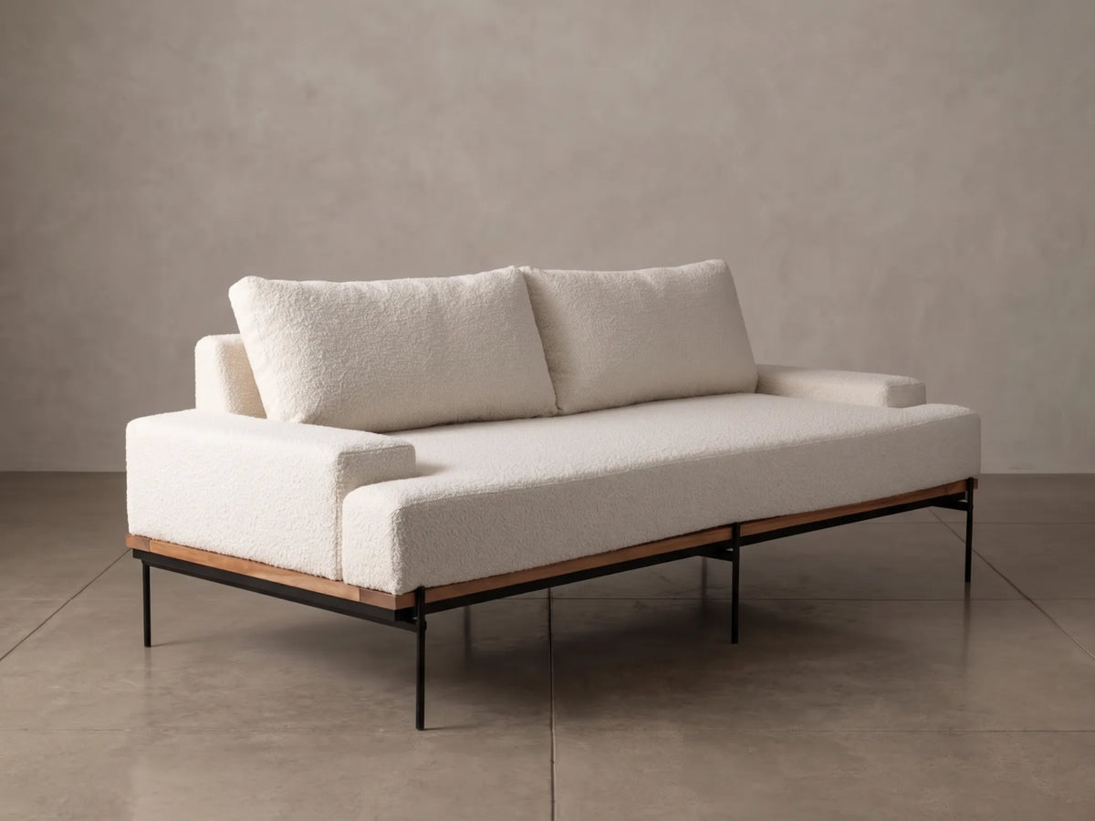 A modern low-profile daybed with textured cream bouclé upholstery, two matching back cushions, a light wood frame, and black metal legs, set against a neutral concrete wall and floor.