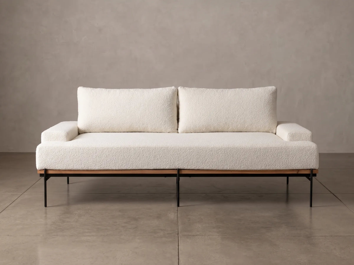 A modern two-seat sofa with textured cream bouclé upholstery, two matching back cushions, and a black metal frame, set against a neutral concrete wall and floor.