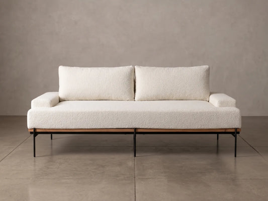 A modern two-seat sofa with textured cream bouclé upholstery, two matching back cushions, and a black metal frame, set against a neutral concrete wall and floor.