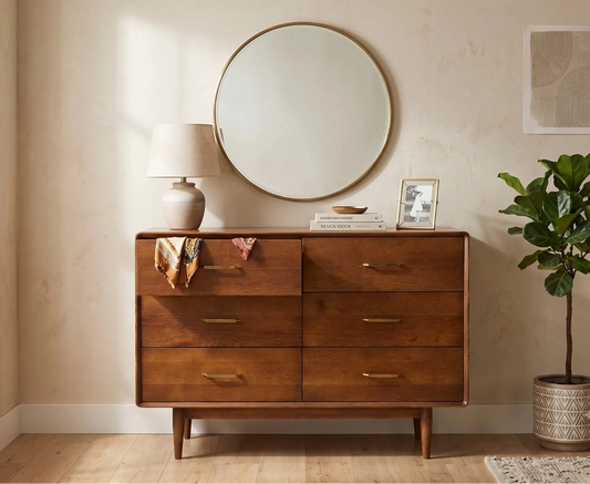 A mid-century modern wooden dresser with six drawers and brass handles, styled in a warm beige room with a round mirror, lamp, framed picture, and a potted fiddle-leaf fig plant.