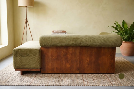 A mid-century modern wooden bench with an olive green velvet upholstered top and side ottoman, styled on a woven rug in a warm, minimalist room.