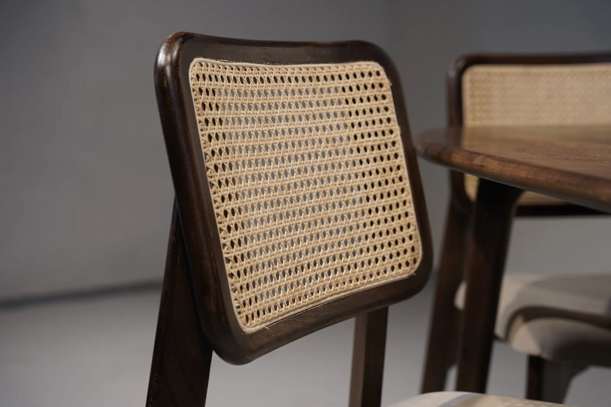 A close-up of a wooden dining chair with a square backrest featuring a woven cane or rattan panel, shown next to a matching wooden table in a softly lit interior.