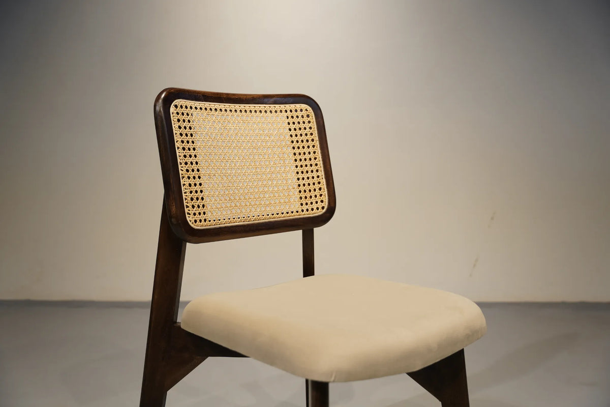 A mid-century modern wooden dining chair with a dark brown frame, a natural cane backrest, and a cream upholstered seat cushion, set against a neutral gray studio background.