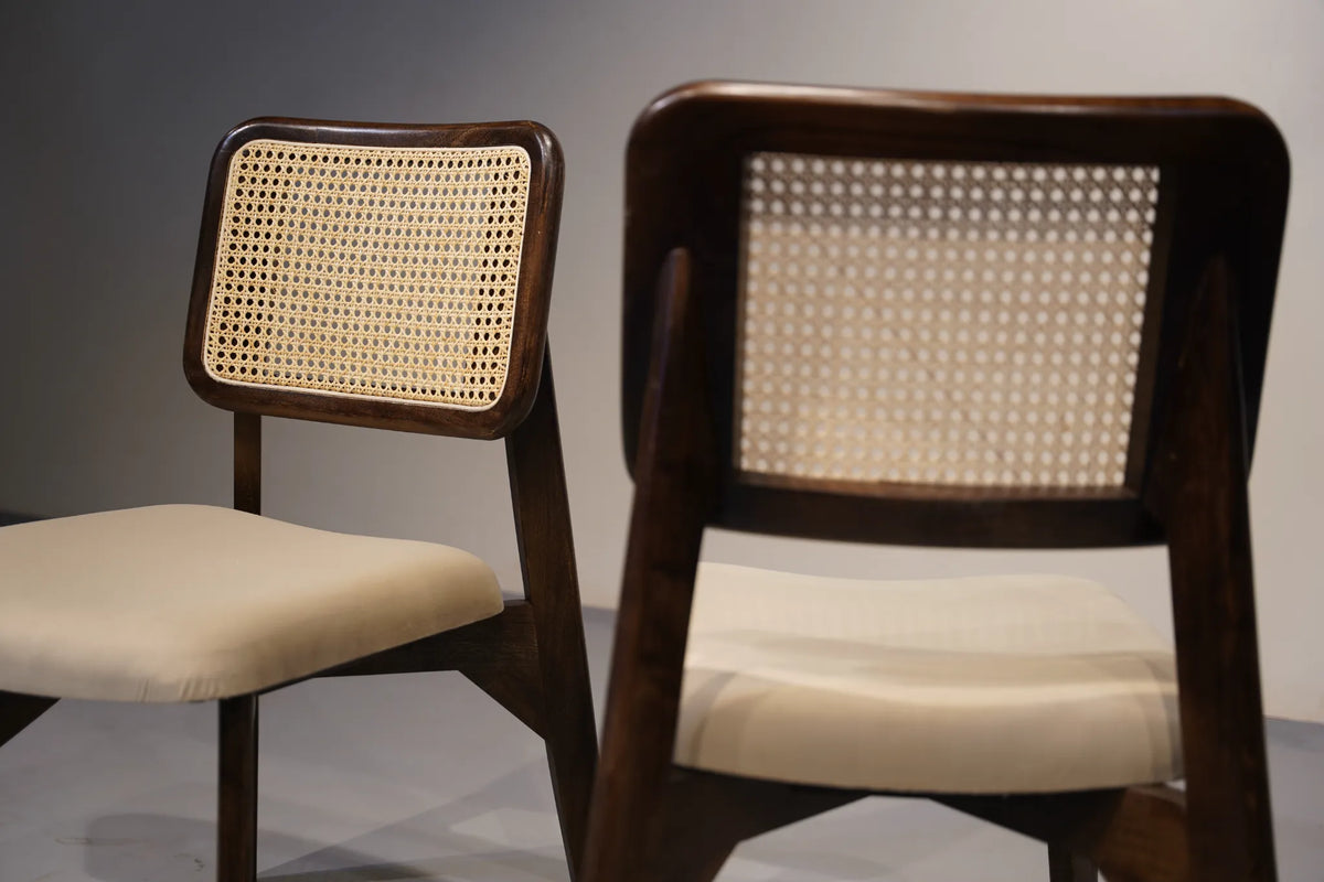 Two mid-century modern wooden dining chairs with woven cane backrests and cream upholstered seats, shown against a neutral gray background.