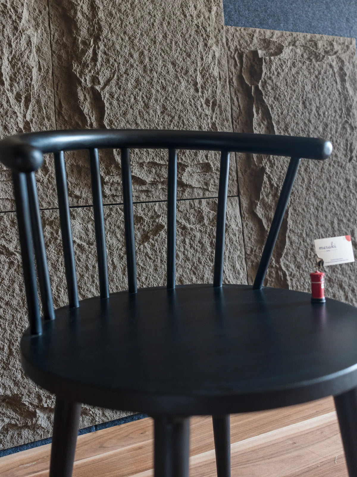 A close up of a black wooden dining chair with a curved backrest and spindle design, set against a textured gray stone wall and light brown wooden floor.