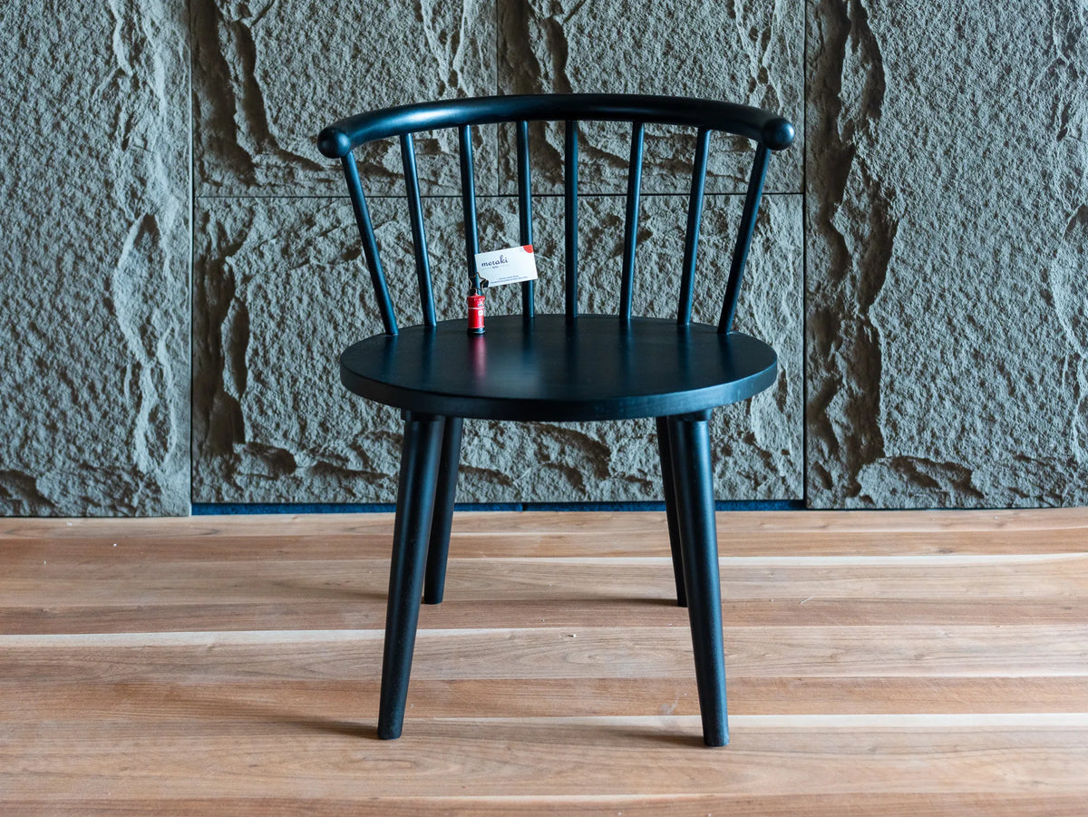 Front view of a black spindle-back chair with a curved backrest and tapered legs, shown on a light wood floor against a textured stone wall with brand name "Meraki Arts"