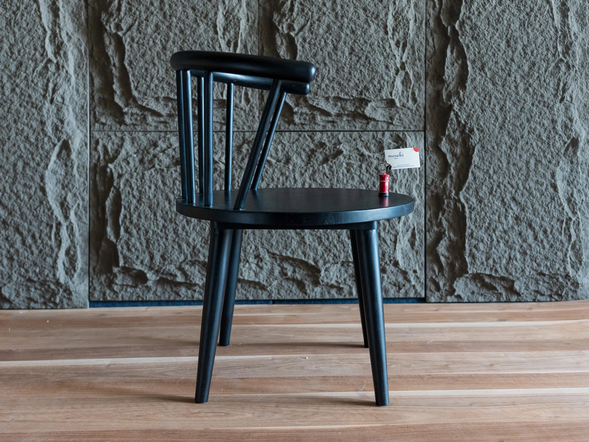 Side view of a black spindle-back chair with a curved backrest and tapered legs, shown on a light wood floor against a textured stone wall.