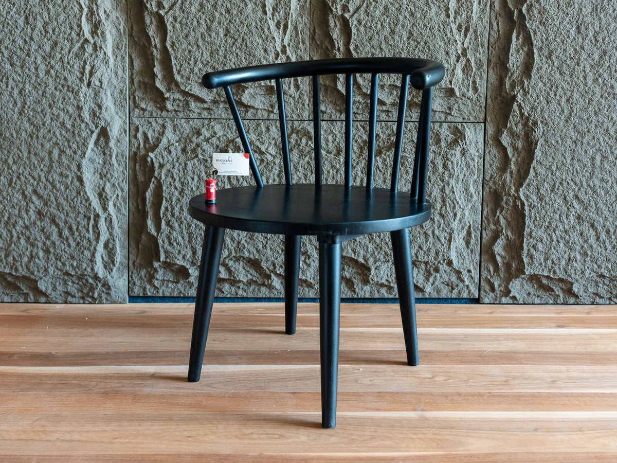 A black spindle-back chair with a curved backrest and tapered legs, shown on a light wood floor against a textured stone wall.